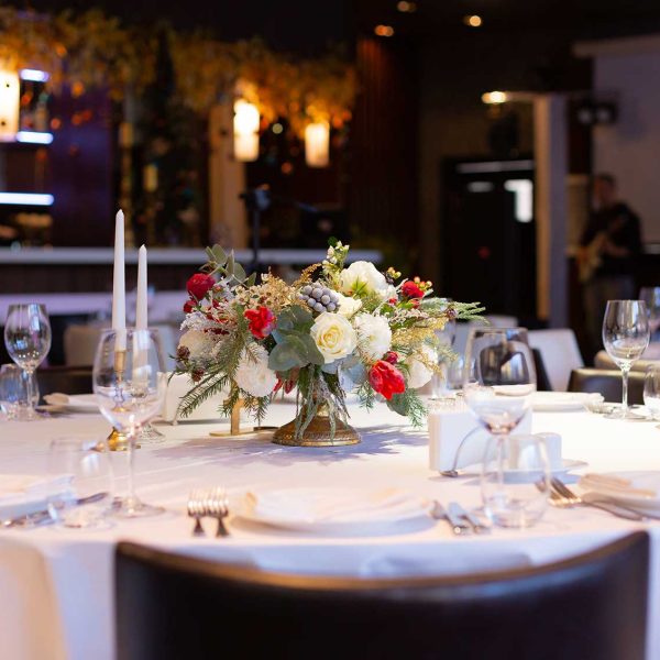 Beautiful spring flowers arrangement with roses, hypericum, tulips standing in a golden vase on a table in a restaurant, for celebrating a birthday, anniversary, wedding. Beautifully served table with glasses, plates, appliances, forks, spoons, a knife, candle napkins in a restaurant. In the background bar and bartender, music band